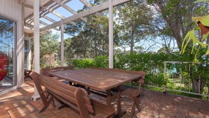 An outdoor patio with a wooden table and benches, surrounded by greenery and a glass enclosure. - Windarra on Denhams Unit Three (Denhams Beach)
