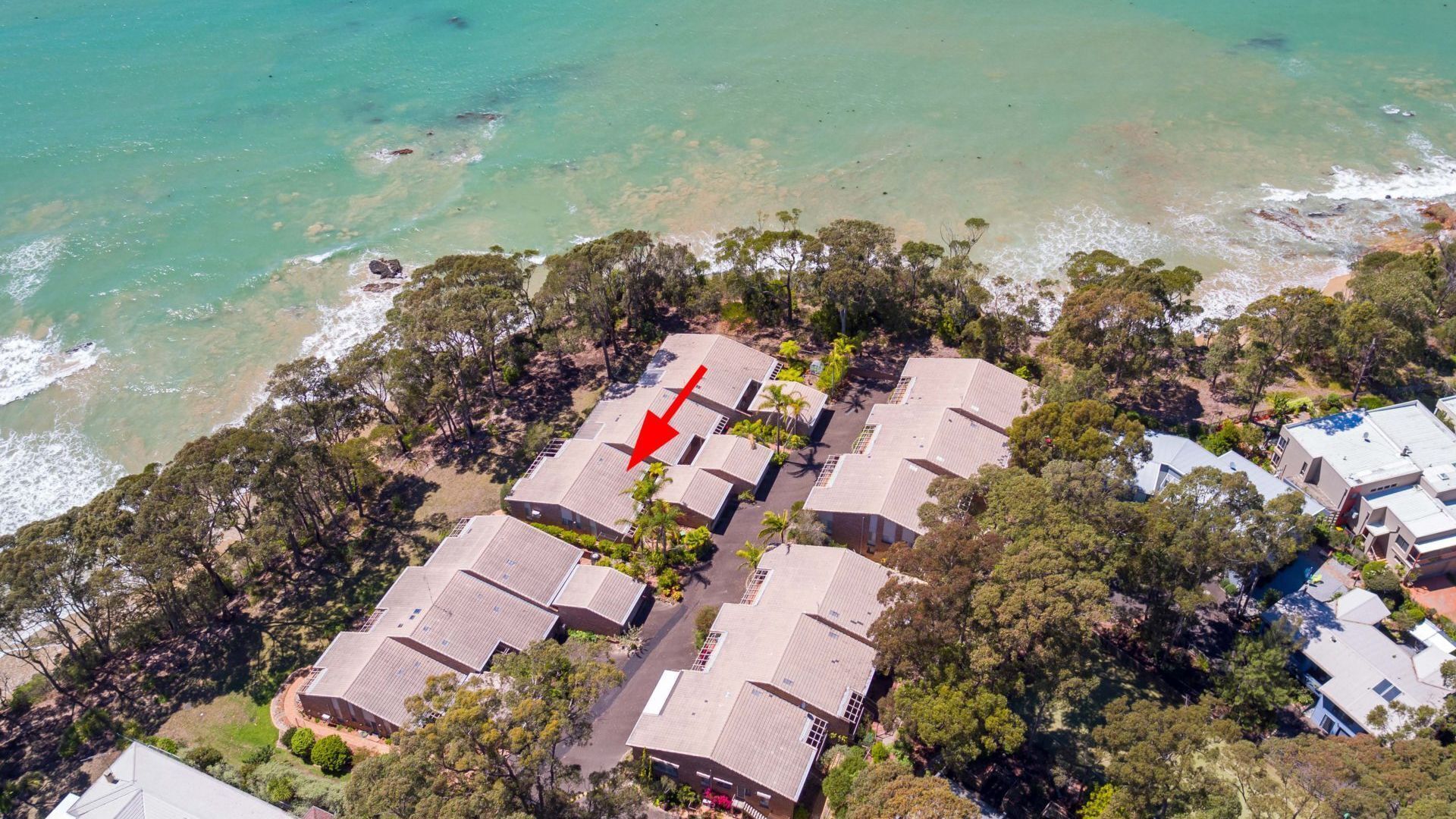 Une vue aérienne d’un quartier résidentiel situé sur la côte, avec de nombreuses maisons, une vue dégagée sur l’océan et une flèche rouge bien visible pointant vers une maison en particulier.