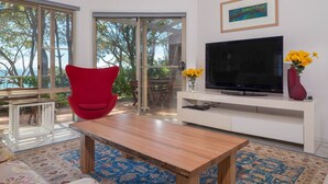 A modern living room with a wooden coffee table, a red chair, a flat-screen TV on a white stand, and a view of trees through large glass doors. - Windarra on Denhams Unit Three (Denhams Beach)