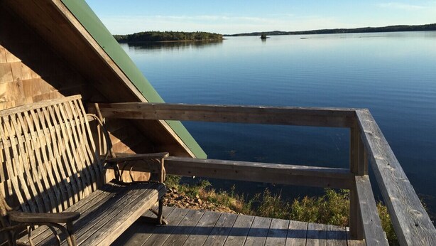 A wooden deck with a bench overlooking a calm lake, a green roof, and a distant shoreline. - Sitting on the edge of Cobscook Bay at Rossport by the Sea (Bali)
