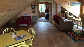 A cozy attic room with wooden walls and a sloped ceiling. It features a dining table with chairs, a sofa, a red chair, and a small cabinet. - Sitting on the edge of Cobscook Bay at Rossport by the Sea (Bali)