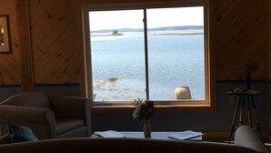 A room with a large window overlooking a body of water, a wooden interior, and a small table with papers and a vase of flowers. - Sitting on the edge of Cobscook Bay at Rossport by the Sea (Bali)