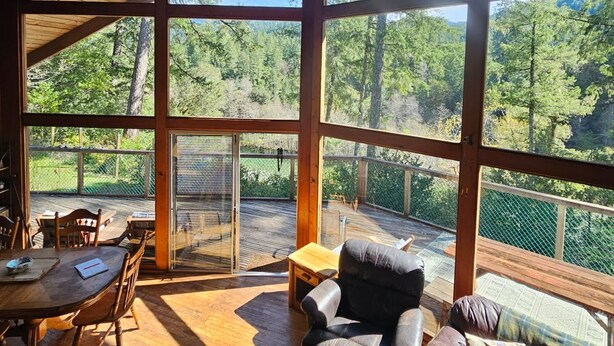 A spacious room with large windows, a wooden ceiling, a dining area, and a seating area with a view of the forest.