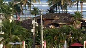 Blick auf einen Strand mit Palmen, ein Gebäude mit Ziegeldach und das Meer im Hintergrund.