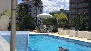 Ein Pool mit Liegestühlen und Sonnenschirmen, umgeben von Palmen und Gebäuden.