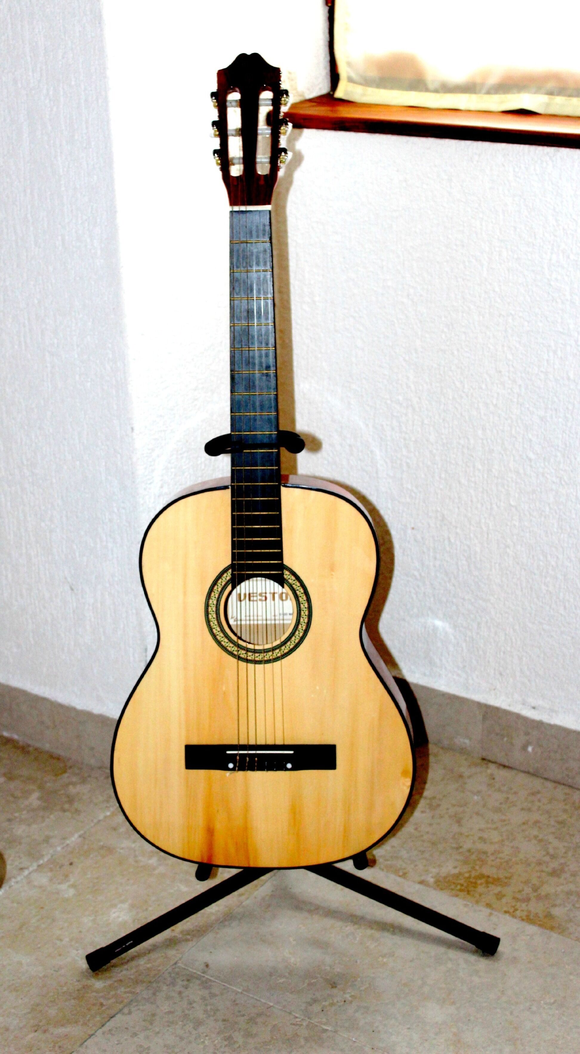 Una guitarra clásica sobre un atril frente a una pared lisa.
