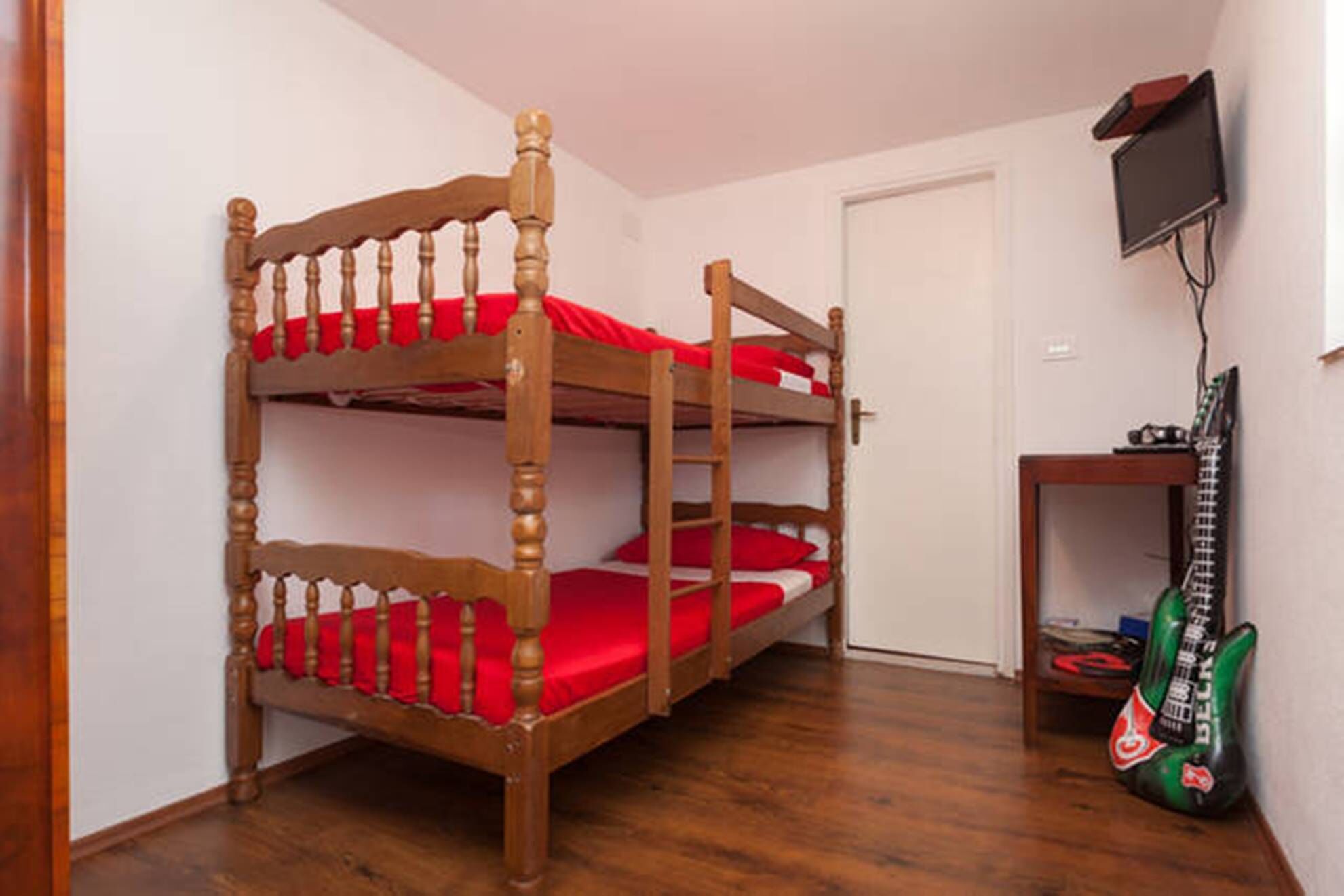 A bunk bed with red bedding in a room with wooden flooring, a door, and a small table with a guitar.