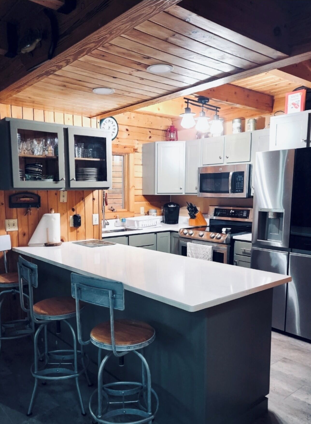Une cuisine avec des murs en bois, des armoires blanches, un îlot central doté de tabourets de bar, des appareils en acier inoxydable et des luminaires suspendus.