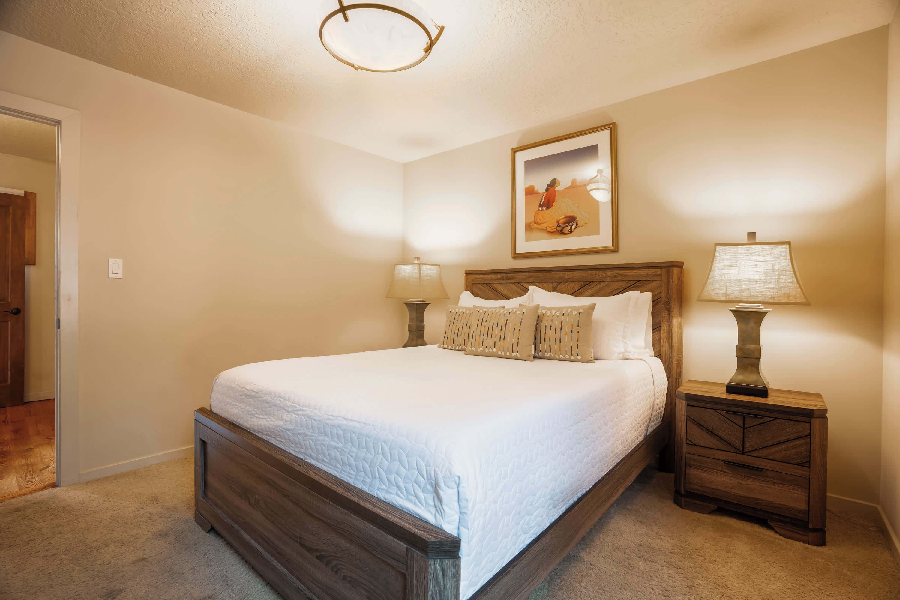 A bedroom with a large bed, two bedside tables with lamps, a framed picture on the wall, and a door to another room.