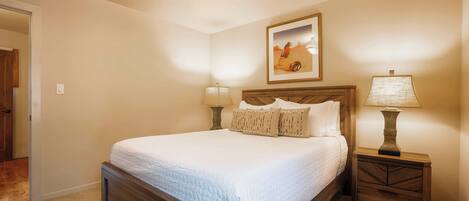A bedroom with a large bed, two bedside tables with lamps, a framed picture on the wall, and a door to another room.