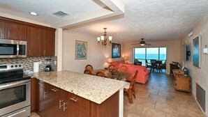 A kitchen with granite countertops, wooden cabinets, and stainless steel appliances. There is a dining area with a table and chairs, and a view of the ocean through large windows.