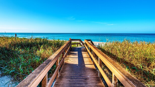A wooden boardwalk leading to a beach with clear blue water and a bright blue sky.