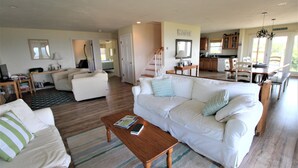 A spacious living room with a white sofa, a wooden coffee table, and a kitchen in the background. - Watch the Sunset from this Bayfront Home! (1349) (Wellfleet)