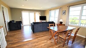 A spacious living area with a wooden dining table and chairs, a dark-colored sofa, a television, and large windows offering a view of the outdoors. - Watch the Sunset from this Bayfront Home! (1349) (Wellfleet)