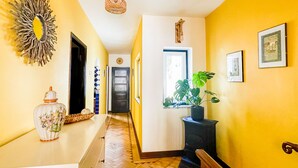 Un pasillo con paredes amarillas, suelo de madera, un espejo decorativo, un jarrón, una planta en maceta y cuadros enmarcados.