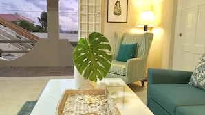 A room with a view of a staircase, a patterned armchair, a teal sofa, a white coffee table with a wicker basket, and a framed leaf artwork. -  Spacious Oceanview One Bedroom Apartment (Atlantic Shores)