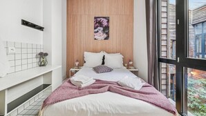 Ein modernes Schlafzimmer mit großem Fenster, einem Holzkopfende, einem Bett mit weißen Leinen, einer pink-weißen Tagesdecke und einer kleinen Vase mit Blumen auf dem Nachttisch.