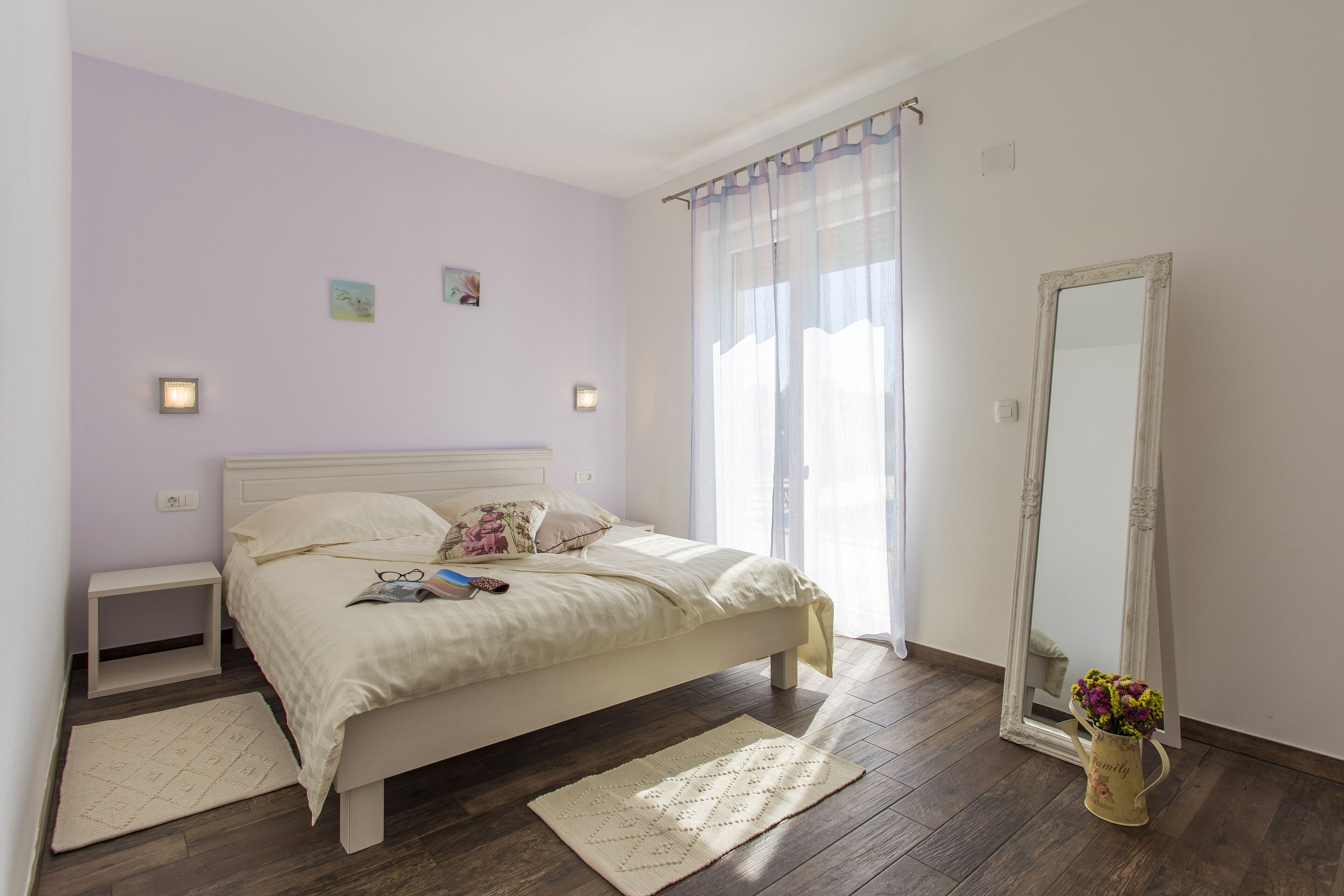 Ein Schlafzimmer mit Bett, Nachttischen, einem Spiegel, einer Vase mit Blumen und einem Fenster mit Vorhängen.