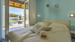 Une chambre à coucher avec un lit, des oreillers et un chapeau posé sur le lit. Il y a une fenêtre donnant sur une piscine et sur du mobilier d’extérieur.