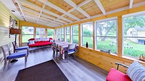 A covered porch with wooden beams, a red couch, a dining table with plaid tablecloths, and large windows offering a view of a yard.