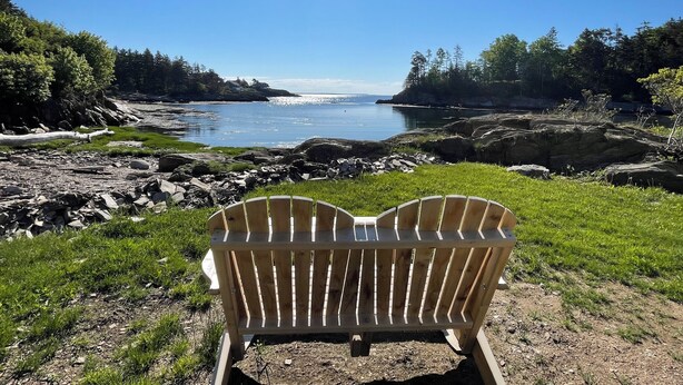 A wooden Adirondack chair facing a rocky shoreline with a clear view of a calm lake and distant trees.