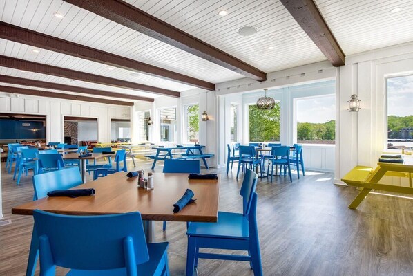 A spacious dining area with blue chairs and wooden tables, large windows offering a view of greenery, and a high ceiling with exposed beams.