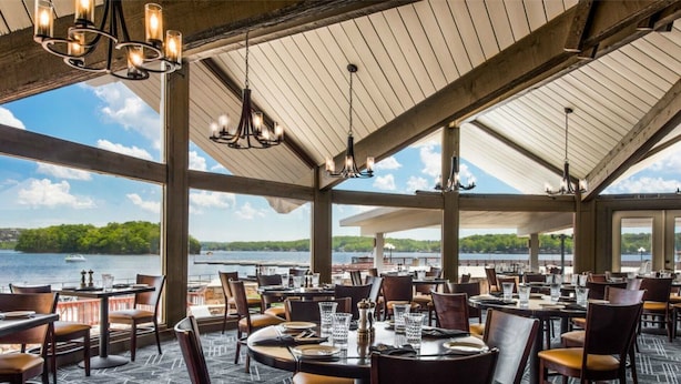 An outdoor dining area with tables and chairs, large windows, and a view of a lake.