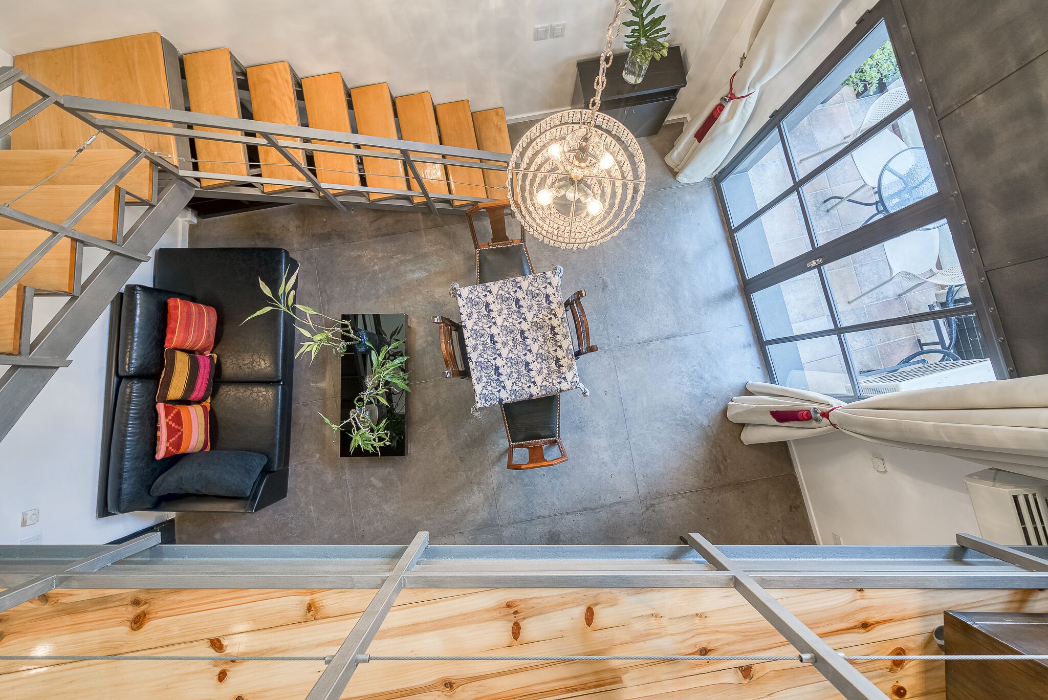 A modern interior with a staircase, a black leather sofa with colorful cushions, a patterned table, and large windows.