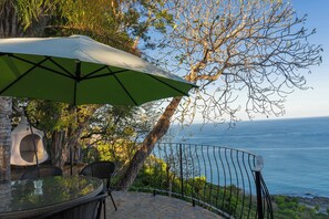 Eine Terrasse mit Glastisch und Stühlen, einem grünen Sonnenschirm und Blick aufs Meer.