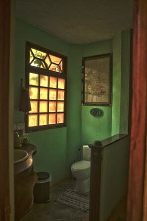 Ein kleines, schmales Badezimmer mit grüner Wand, einem Fenster mit geometrischem Muster, einer Toilette und einem Waschbecken auf Holzfüßen.