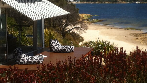 A beach house with a glass enclosure, two outdoor seating areas with polka dot cushions, and a view of the beach and ocean.