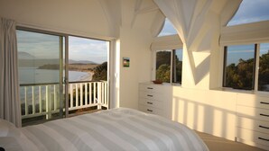A bedroom with a large bed, a sliding glass door to a balcony, a dresser, and a window with a view of the beach and hills.