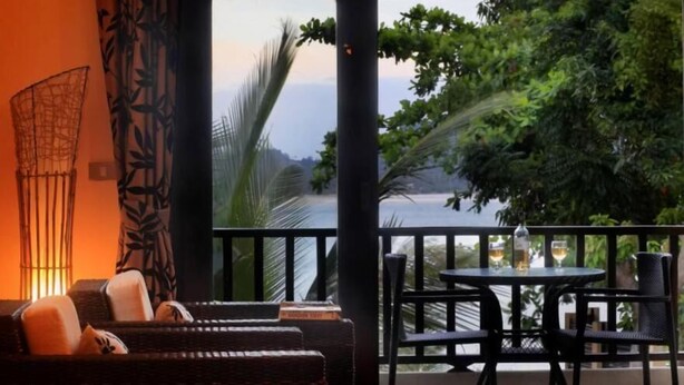 A balcony with wicker furniture, a table with drinks, and a view of a body of water and greenery.