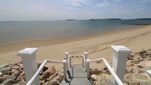 Une plage avec un escalier descendant vers l’eau, entourée de rochers et bordée d’un ciel dégagé.