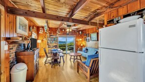 L’intérieur chaleureux d’un chalet avec un plafond en bois, un coin repas comprenant une table et des chaises, un canapé, un réfrigérateur et une vue sur l’extérieur grâce à une porte vitrée.