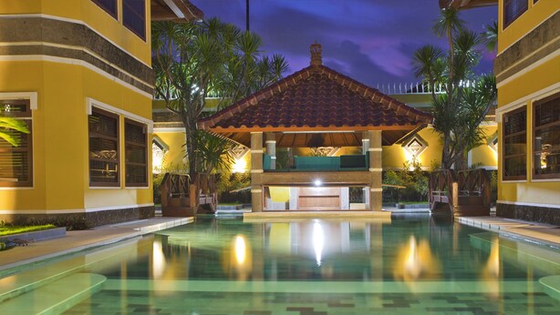 A swimming pool with a covered area, palm trees, and a building with yellow walls and a tiled roof. - Sanur, Elegant 2 Bed Villa
 (Sanur)
