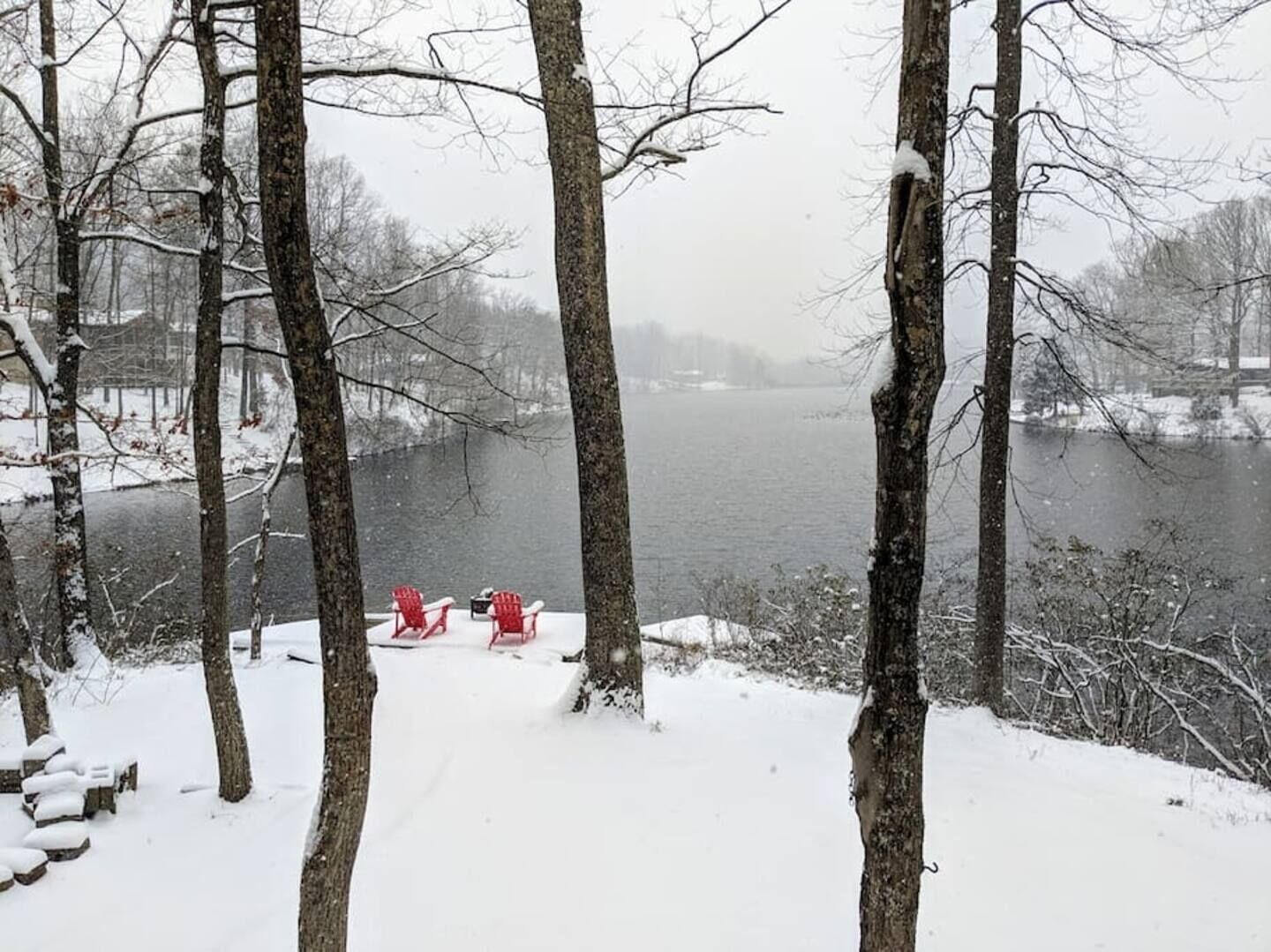 Paisaje nevado con un lago, árboles y dos sillas Adirondack rojas.