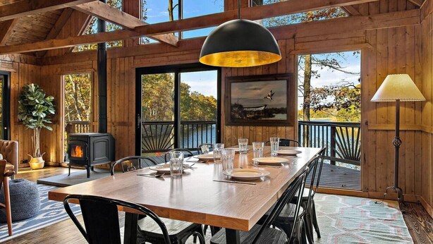 A wooden interior with a dining table set for four, a fireplace, and large windows offering a view of the outdoors.