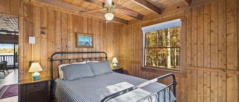 A wooden cabin bedroom with a bed, two bedside tables, a ceiling fan, and a window with a view of trees.