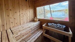 A wooden sauna with a bench, a window with a view of trees and a lake, and a container with rocks.