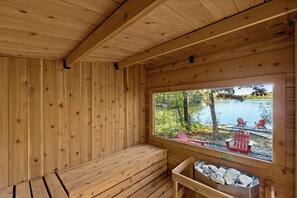 A wooden sauna with a bench and a window overlooking a lake with chairs outside. - Cozy Cabin | Lakefront, Hot Tub, Outdoor Sauna (Harpers Ferry)