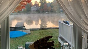 Blick durch ein Fenster mit weißen Vorhängen, ein Garten mit Teich, einem blauen Pool und einer Holzterrasse.