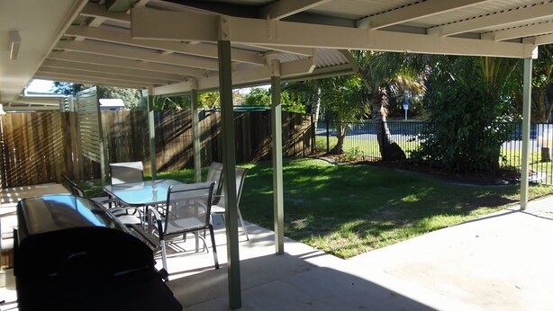 Un patio techado con una mesa, sillas, una parrilla y vistas a un jardín con césped y árboles.