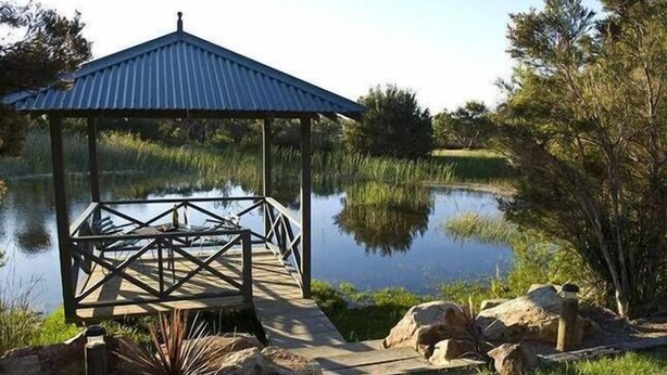 A gazebo with a metal roof and wooden floor, surrounded by rocks and grass near a calm pond. - Howards Hill bed & breakfast (cape schanck)