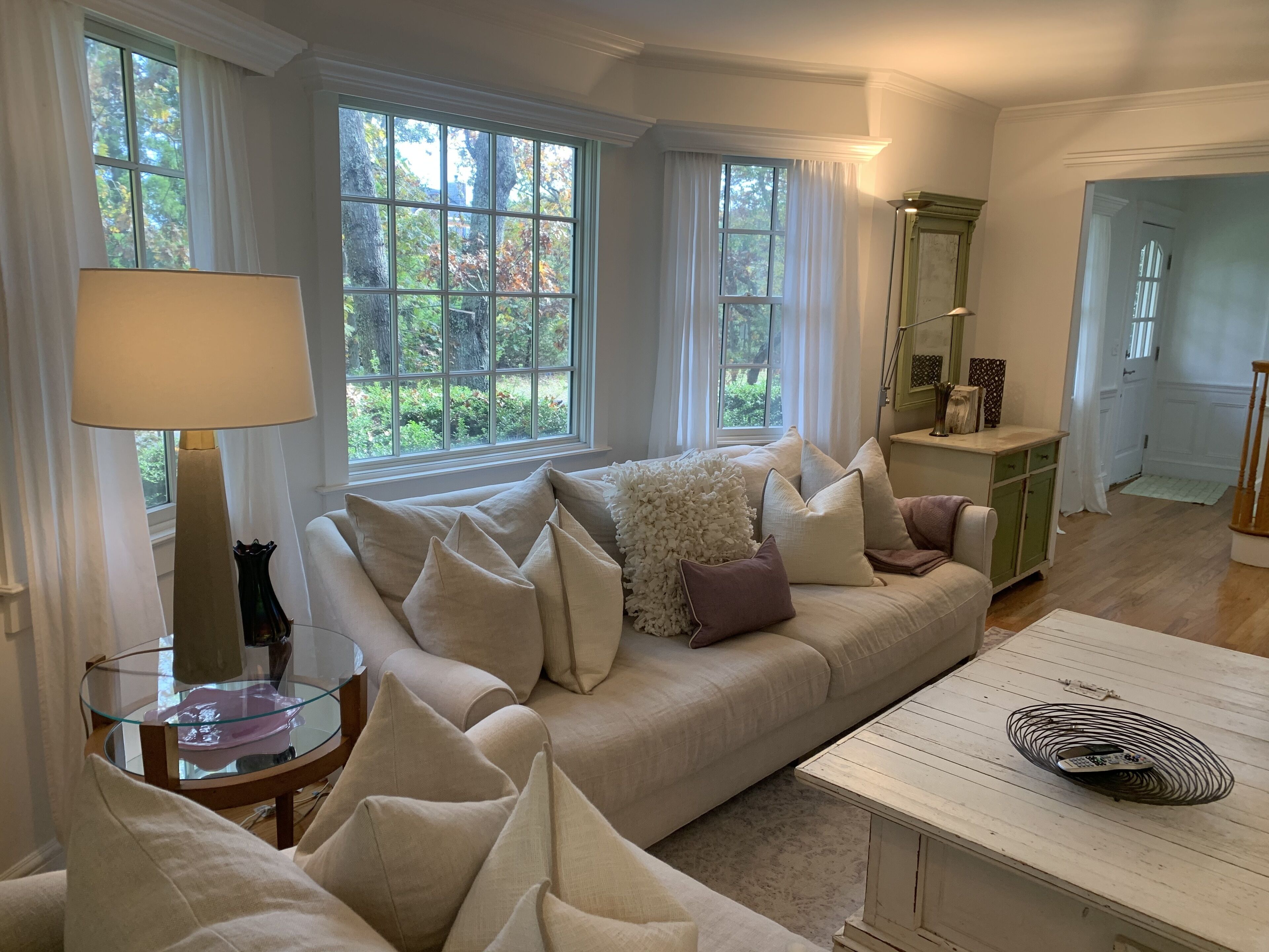 A cozy living room with a beige sofa, a glass coffee table, and a lamp. There are two windows with curtains, a green cabinet, and a door leading to another room.