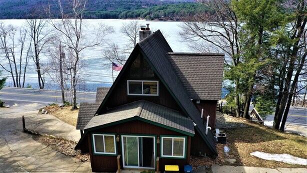 A small, two-story cabin with a steep gabled roof, located near a body of water and surrounded by trees. - Great Sacandaga Lakefront Home with 100 feet of deep water access (Hadley)