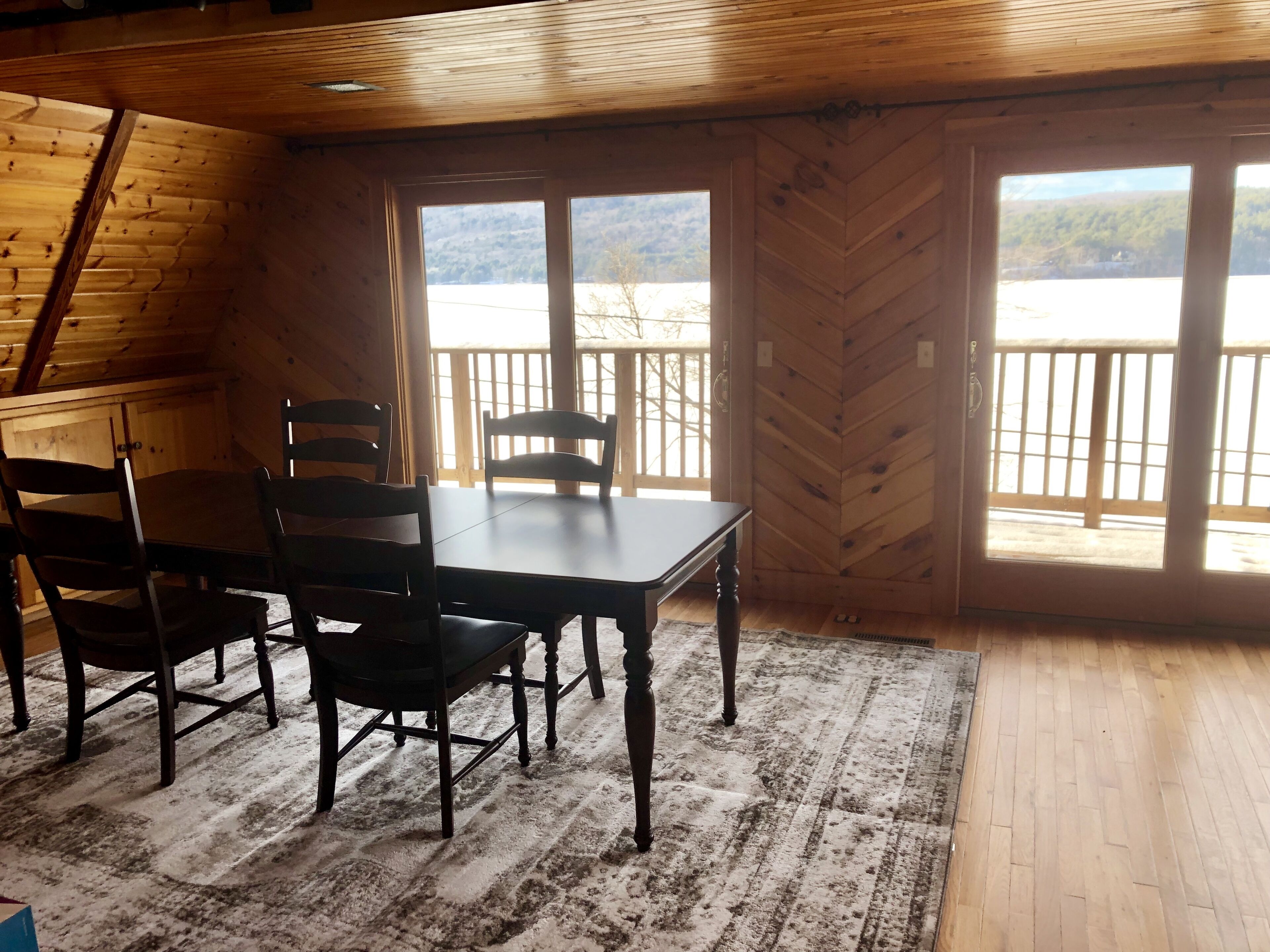 Una cabaña de madera con comedor, que incluye una mesa, sillas, un revestimiento de pared con motivo de espiga y ventanas amplias con vistas a un lago y un bosque.
