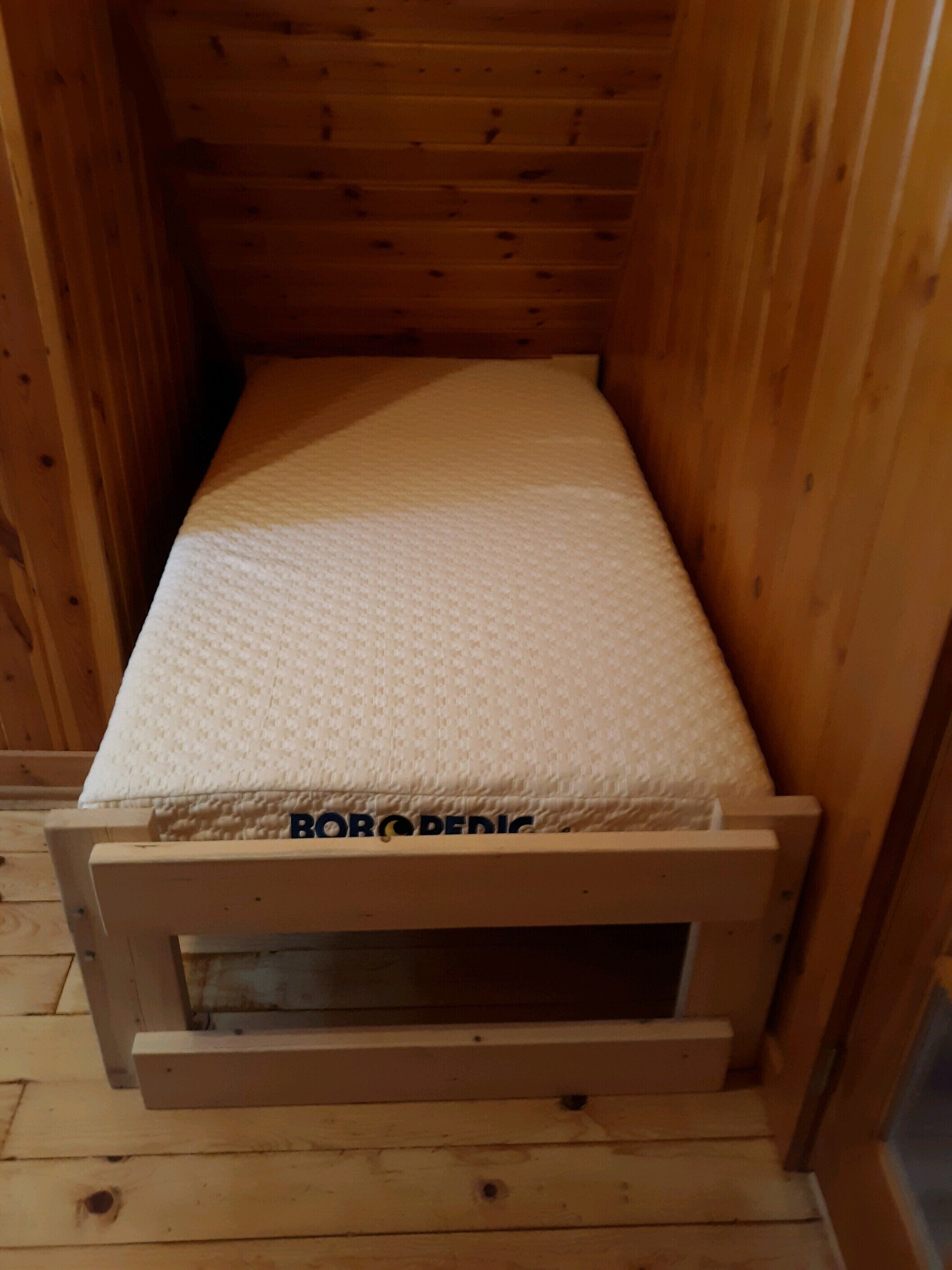 Una cama pequeña con estructura de madera y colchón blanco en una habitación tipo cabaña.