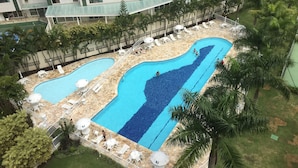 A swimming pool with two separate sections, surrounded by lounge chairs and umbrellas, with palm trees and a building in the background. - 🌺RIVIERA APARTMENT WITH SEA VIEW (send message before) (Bertioga)