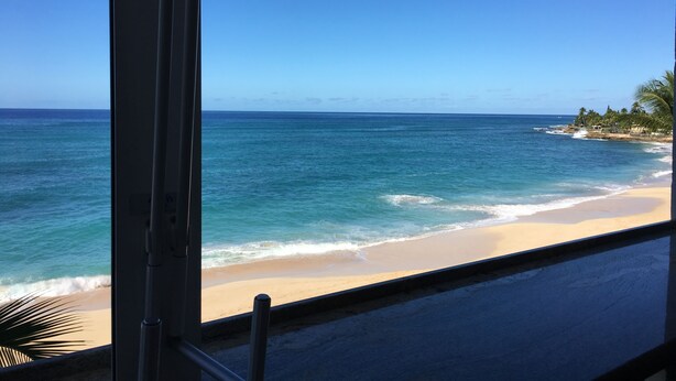 A beach view from a window with a clear sky, ocean, and a sandy shore with palm trees. - Cozy Beach front condo in Makaha (Waianae)